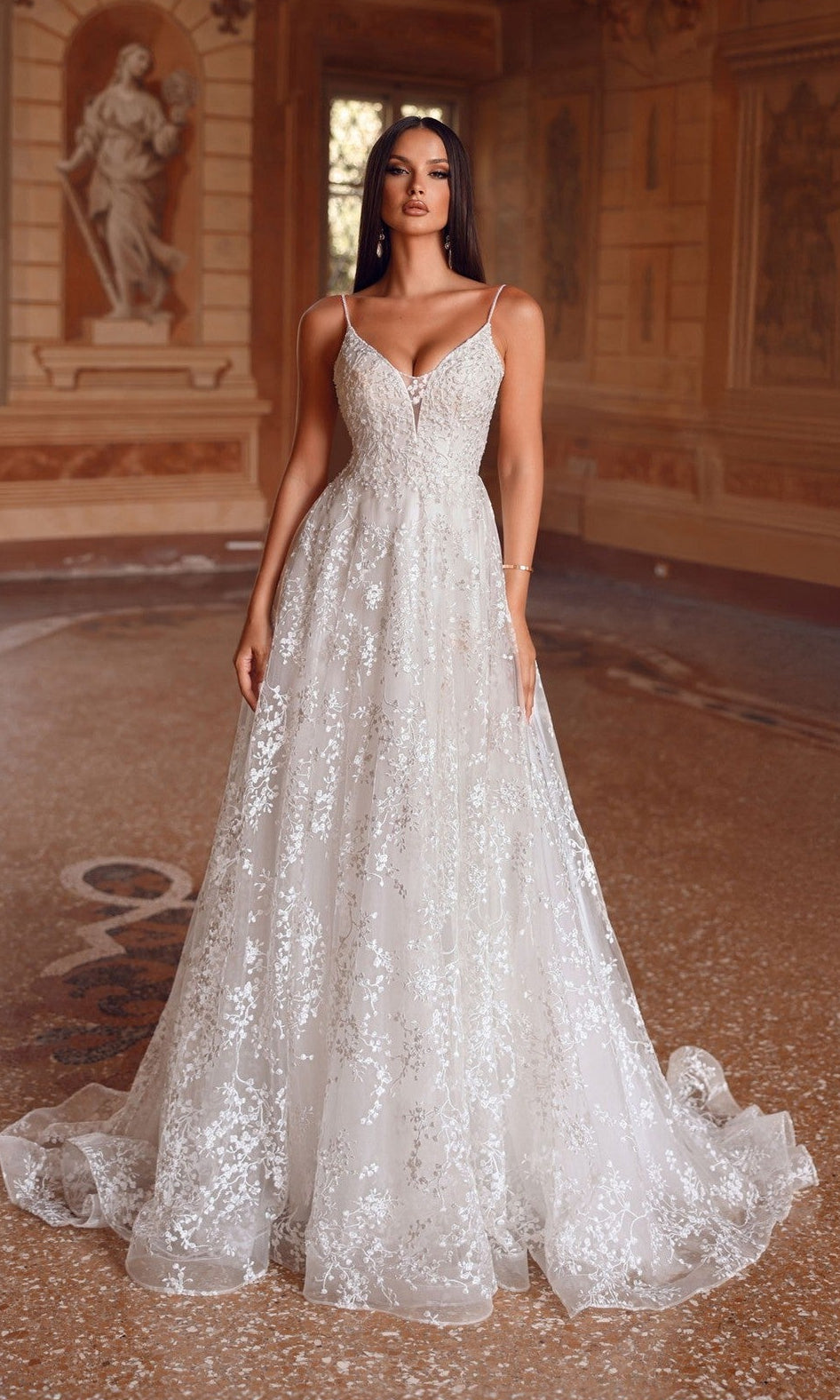 A woman stands in a grand hall wearing the Alyce 7121 long ivory dress, featuring thin straps, a fitted bodice, and delicate floral lace details.